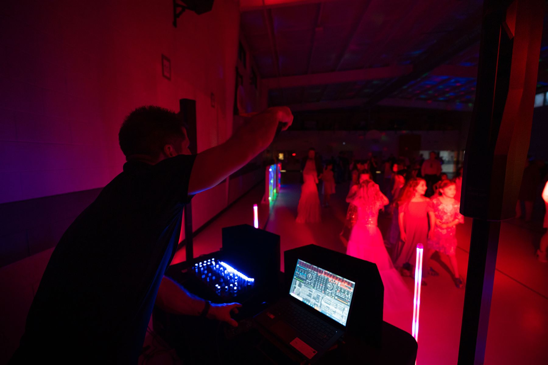 A DJ at a laptop and lights, with a school gym glowing red and blue behind him