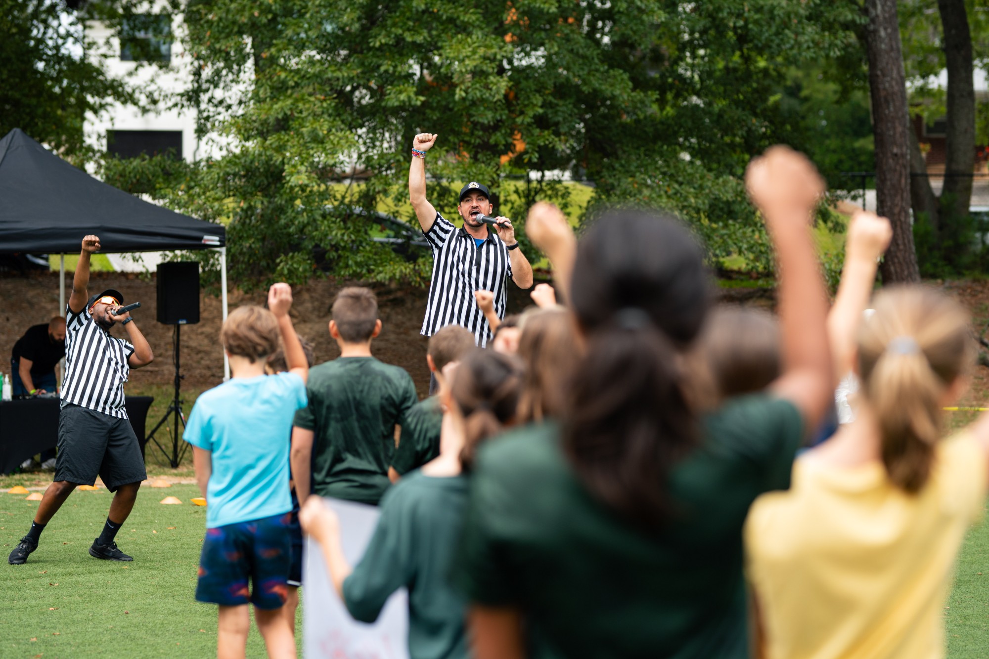 Two emcees in referee shirts with microphones and raised fists leading a crowd of students outdoors