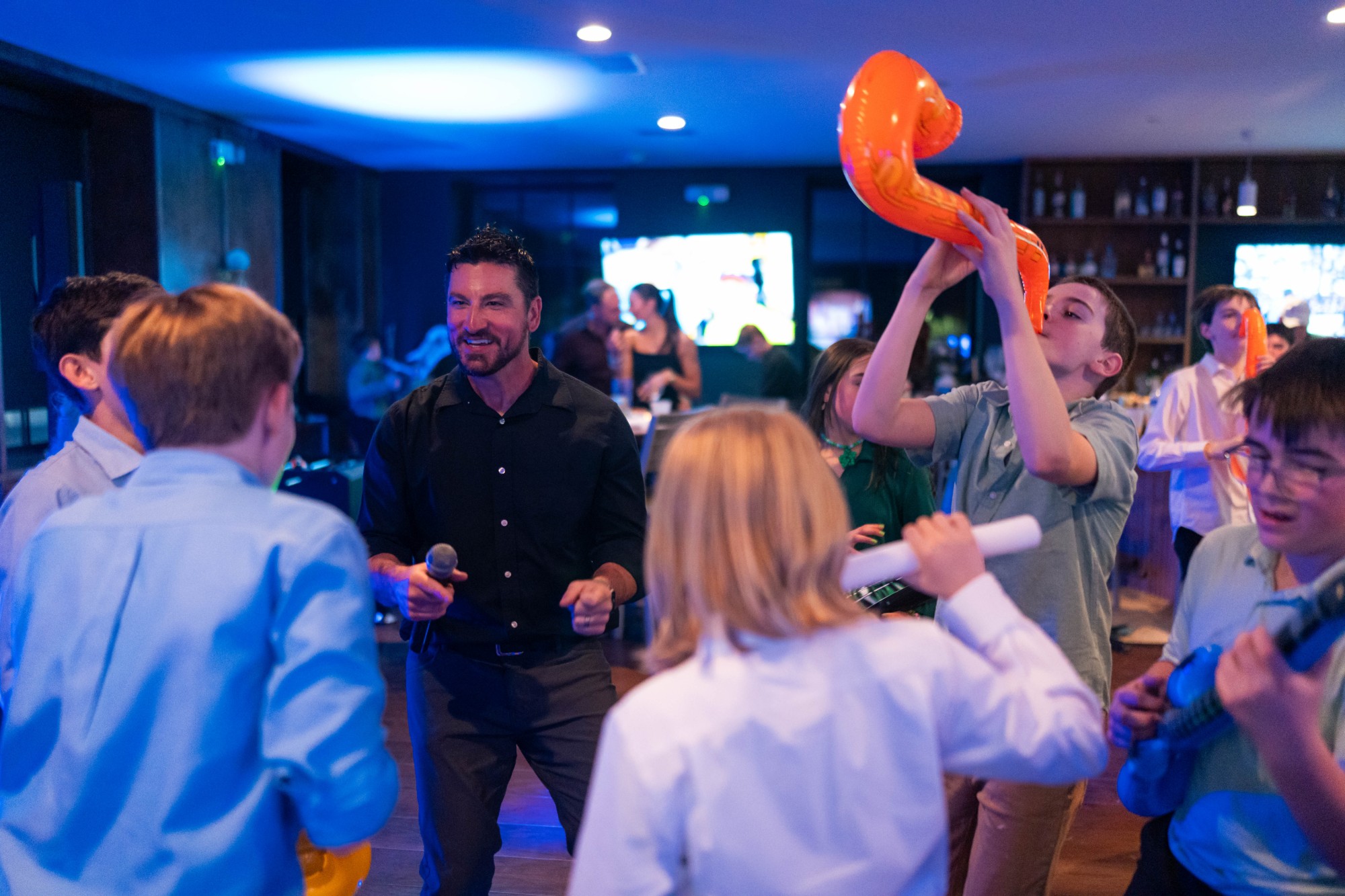 Brian smiling with a microphone in the middle of kids playing inflatable instruments on the dance floor